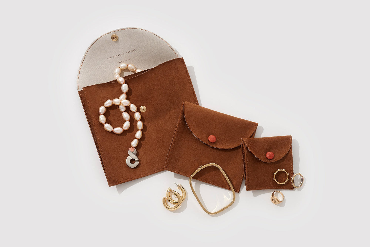 Three brown pouches with red buttons, a pearl necklace, gold hoop earrings, a square bangle, and two rings on a light background.