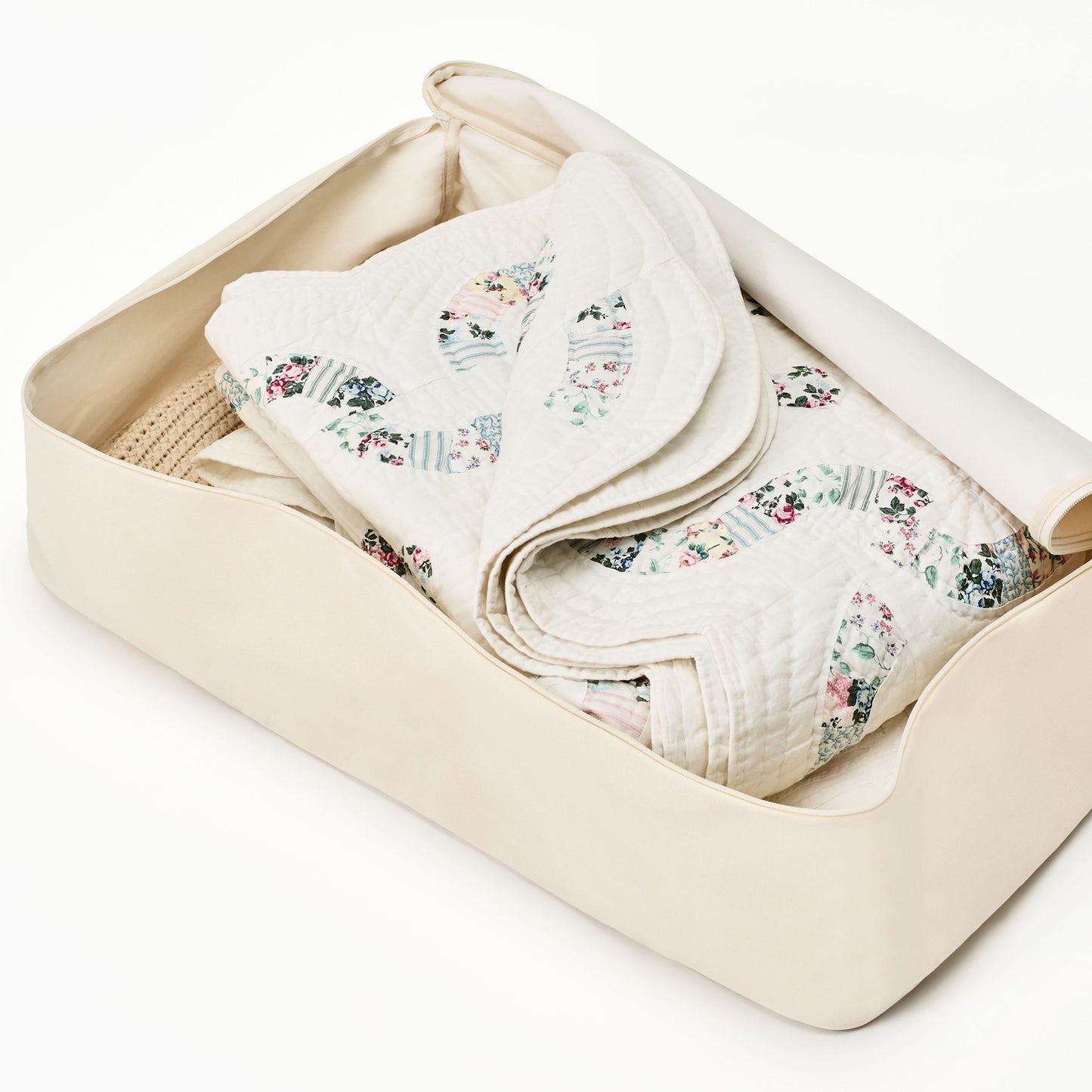 A folded quilt with a floral pattern is partially tucked inside a beige storage bag on a white background.