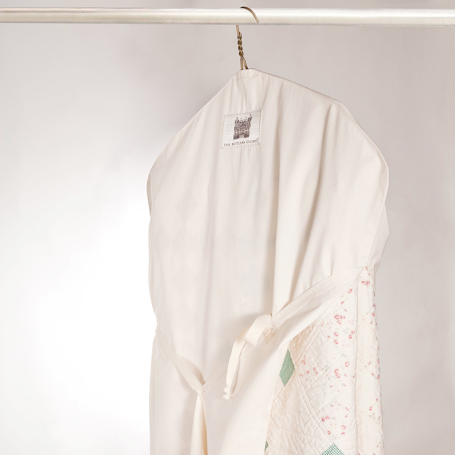 A white garment bag hangs on a rod, containing a quilt with floral and green geometric patterns.