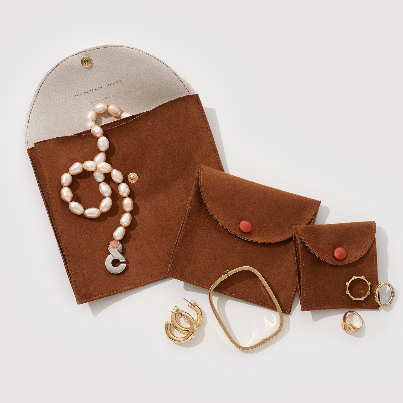 Jewelry including pearl necklace, rings, and earrings placed beside three brown pouches with red buttons.