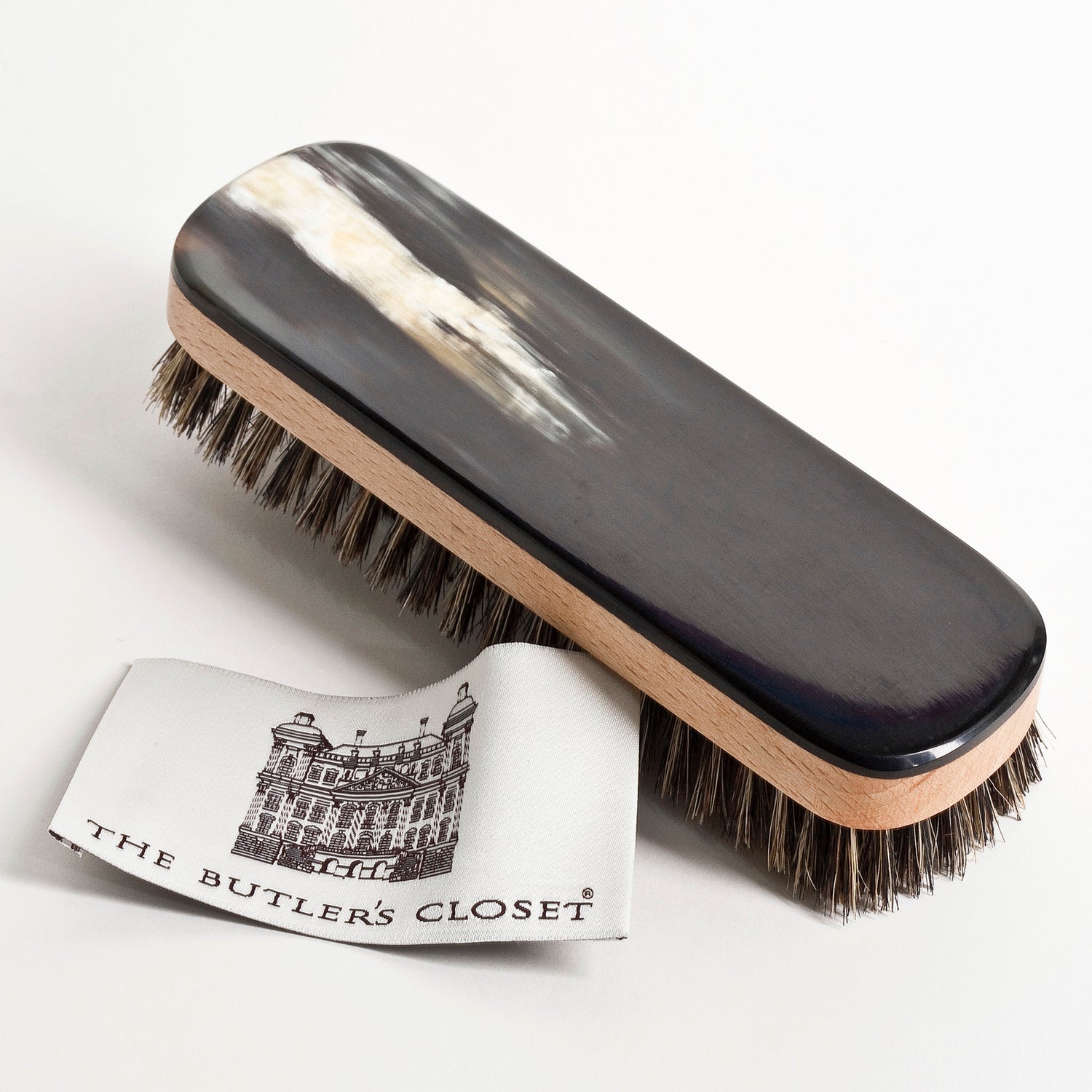 A shoe brush with a dark wooden handle and bristles is placed next to a white tag labeled The Butler's Closet.