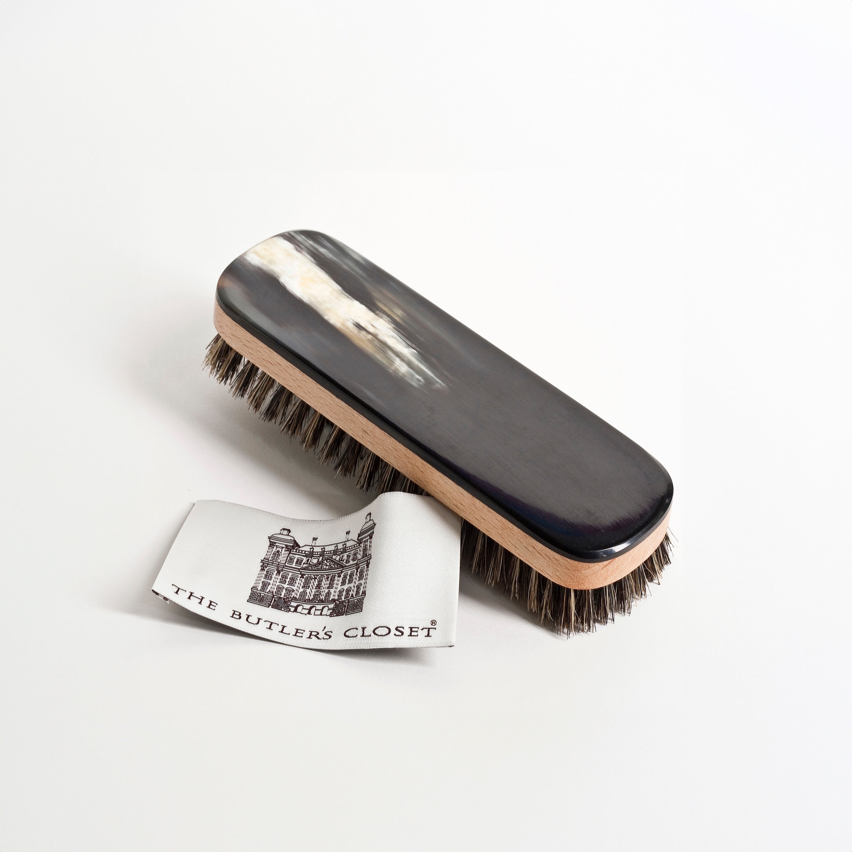 A clothes brush with mixed bristles and a dark, polished wooden handle alongside a small card featuring a building illustration and the text The Butler's Closet.