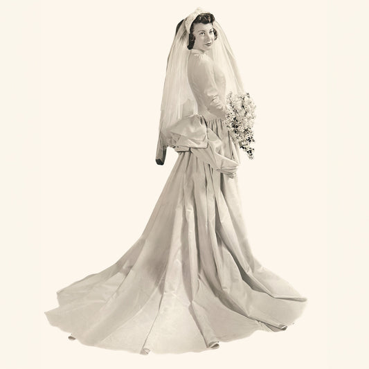 A bride in a vintage gown poses with a bouquet, wearing a long veil and holding flowers.